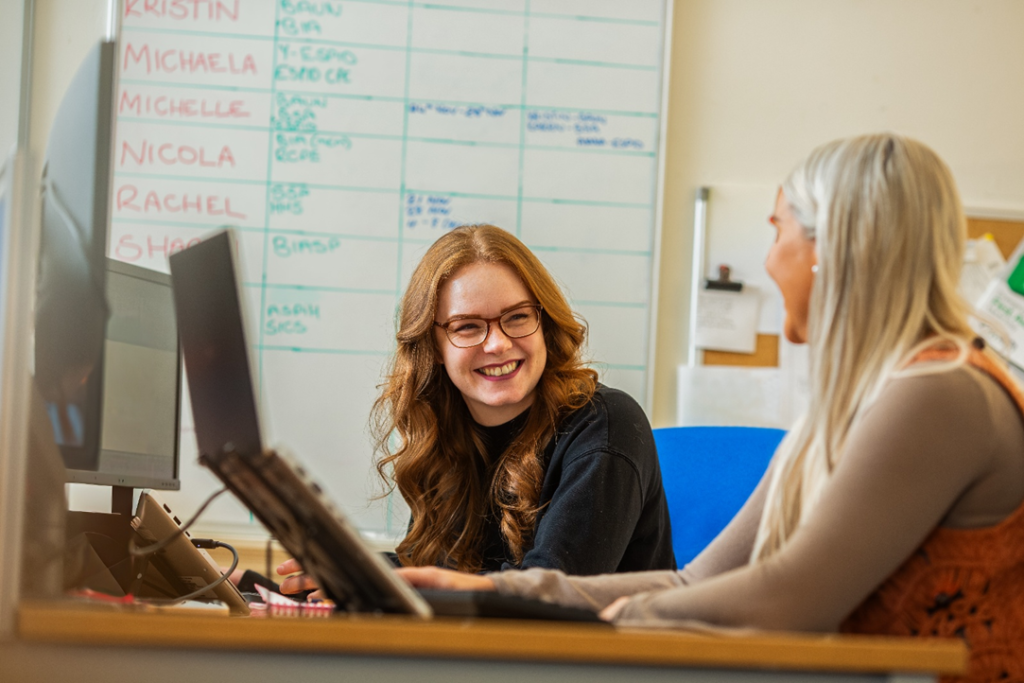 Two office staff working together at their computers, sharing a light moment and laughing. The scene shows a positive, collaborative workplace environment with teamwork, communication, and a friendly office culture in a modern workspace.