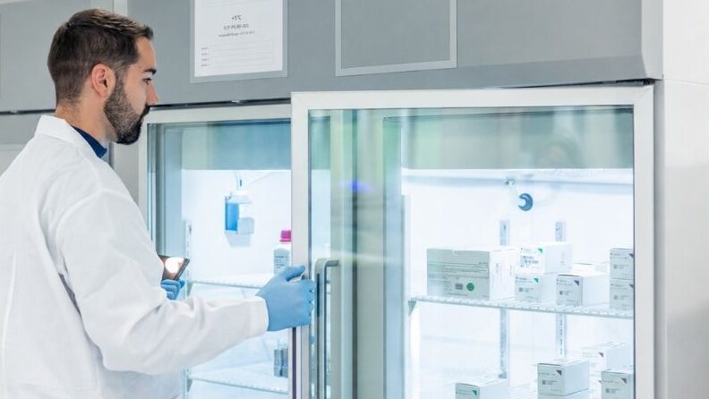 A healthcare professional in a white coat and blue gloves carefully checks medical supplies inside a glass-fronted refrigerated storage unit.